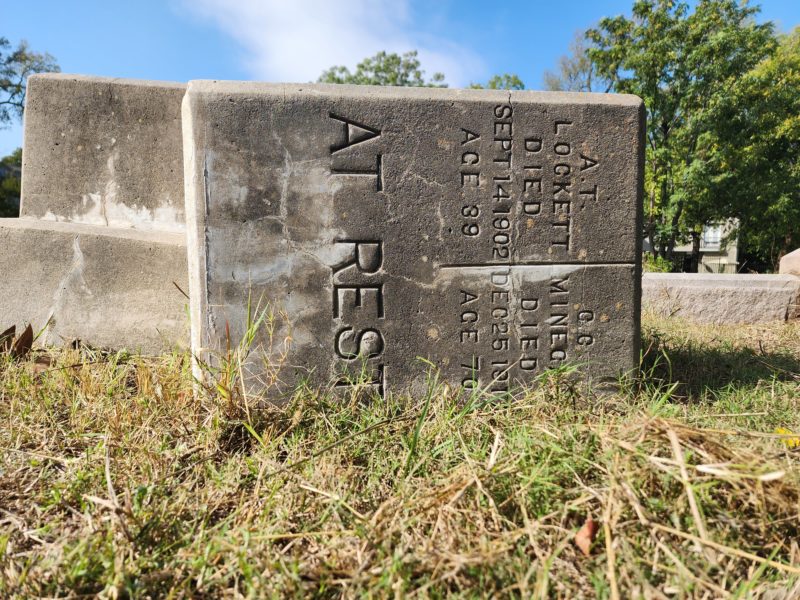 A gray cement headstone in the shape of a rectangle is laying on its side in the grass. On the front is the inscriptions for two people. The one on the left is for A.T. Lockett. The one on the right side are the deatails for C.C. Minegan.